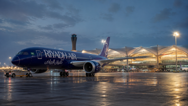 A Riyadh Air commercial aircraft parked on the tarmac at King Khalid International Airport under a modern lit terminal roof at dusk.